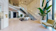 Located at 15 Washington Ave in Suffern, NY, this grand residential lobby delivers an immediately striking first impression through its double-height ceilings and expansive floor-to-ceiling marble wall cladding with warm gold veining. The centerpiece of the space is a dramatic floating staircase with sleek black metal framing and glass balustrades, ascending alongside the soaring marble feature wall. Large floor-to-ceiling windows flood the interior with abundant natural light, offering open views of the surrounding exterior. The main lobby area is finished with large-format light tile flooring and anchored by a stylish lounge seating arrangement, complete with a sofa, accent chairs, and a patterned area rug. Ambient wall sconces and a sculptural chandelier contribute to the refined atmosphere. A secondary lounge area transitions into a more intimate residents' amenity space, featuring bold graphic wallcovering, built-in banquette seating, lounge chairs, and a beverage station framed by dark cabinetry and pendant lighting. Additional functional elements include a mailroom corridor with individual resident mailboxes, slatted wood column accents, and curated artwork throughout. Elevator access and lush indoor greenery complete this polished, upscale communal environment.