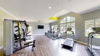 Located in Hoover, Alabama, this well-appointed fitness center offers a comprehensive and energizing workout environment. The space is defined by a clean, modern aesthetic, featuring light hardwood-style flooring, soft gray walls, and bold yellow accent walls that add a vibrant, motivating pop of color throughout.

The cardio zone is positioned along a wall of large, floor-to-ceiling windows that flood the room with natural light and provide pleasant outdoor views, creating an open and airy atmosphere. Multiple treadmills and elliptical machines are thoughtfully arranged in this area, complemented by a wall-mounted flat-screen television. Ceiling fans overhead help maintain comfortable airflow during workouts.

On the opposite side of the room, a dedicated strength training area features a multi-station cable machine, an adjustable weight bench, and a well-stocked dumbbell rack with a variety of free weights, as well as a selection of medicine balls. A large wall mirror in this zone enhances the sense of space and allows users to monitor their form. Crown molding and recessed lighting add a refined architectural touch throughout.

French doors connect the fitness room to an adjacent area, and a secondary exit door provides additional access to the outdoors. The overall layout is spacious and well-organized, catering to a full range of fitness needs in a bright, functional setting.