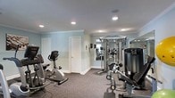 Located at 5055 Harbor Lake Drive in Goose Creek, South Carolina, this fitness center offers a dedicated space for exercise and wellness. The room features soft blue-toned walls accented with crown molding, creating a clean and calm atmosphere that complements an active lifestyle. Recessed lighting throughout the ceiling provides bright, even illumination across the entire workout area. The space is outfitted with a range of exercise equipment, including a stationary recumbent bike, a multi-function cable and weight machine, and additional strength and cardio machines. A large wall mirror on one side of the room enhances the sense of space and allows users to monitor their form during workouts. The durable, textured carpet flooring provides cushioning and stability underfoot, ideal for a fitness environment. A private door leads to an adjoining area, adding to the functionality of the space. The overall layout is thoughtfully organized to accommodate multiple users and various types of training, making this fitness center a well-appointed and practical amenity.