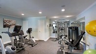 Located at 5055 Harbor Lake Drive in Goose Creek, South Carolina, this fitness center offers a dedicated space for exercise and wellness. The room features soft blue-toned walls accented with crown molding, creating a clean and calm atmosphere that complements an active lifestyle. Recessed lighting throughout the ceiling provides bright, even illumination across the entire workout area. The space is outfitted with a range of exercise equipment, including a stationary recumbent bike, a multi-function cable and weight machine, and additional strength and cardio machines. A large wall mirror on one side of the room enhances the sense of space and allows users to monitor their form during workouts. The durable, textured carpet flooring provides cushioning and stability underfoot, ideal for a fitness environment. A private door leads to an adjoining area, adding to the functionality of the space. The overall layout is thoughtfully organized to accommodate multiple users and various types of training, making this fitness center a well-appointed and practical amenity.
