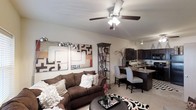 This single-story residence showcases an open-concept layout that connects the living, dining, and kitchen areas into one cohesive and functional space. The living area is anchored by a plush, dark brown sofa accented with decorative pillows, positioned beneath a ceiling fan with integrated lighting that enhances both comfort and illumination. A bold, multi-panel abstract artwork adorns the wall, adding a contemporary artistic touch, while a tall corner display shelf provides additional decorative storage.

The dining area transitions naturally from the living space, featuring patterned upholstered chairs arranged around a dark-toned table, creating an intimate setting ideal for everyday meals or entertaining. Adjacent to the dining zone, the kitchen is outfitted with dark cabinetry, a contrasting countertop with bar seating, and track lighting overhead, providing both style and practicality. The consistent use of dark furniture against light-toned walls and flooring creates a striking visual contrast throughout. Light-colored carpet underfoot and neutral wall tones contribute to a bright, welcoming ambiance across the entire open floor plan.