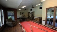 This single-floor recreational space is designed to serve as a dedicated game room and social hub. The room is generously sized, accommodating multiple game stations without feeling cramped. At the forefront is a full-sized pool table with a vibrant red felt surface, complete with cues ready for use. Behind it sits a regulation ping pong table, and to the right, a classic foosball table rounds out the gaming options. Two ceiling fans are mounted overhead, promoting air circulation throughout the space, while fluorescent lighting ensures the room is well-lit for gameplay. The flooring consists of dark hardwood, lending a warm, grounded aesthetic to the interior. Walls feature a two-tone design with a dark wainscoting detail along the lower portion, accented by neutral upper walls. Decorative wall art and a wagon wheel accent piece add character. A wall-mounted television is visible near the entrance area, and a window along one wall allows natural light to filter in. Seating, including a cushioned armchair, provides a comfortable spot for spectators or those taking a break between games.