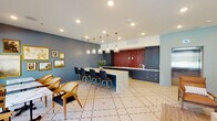 This thoughtfully designed single-floor space blends mid-century modern aesthetics with contemporary finishes to create a versatile and visually striking environment. The open layout is anchored by a generously sized kitchen and bar area, where dark navy cabinetry with brass hardware frames a bold, glossy red brick accent wall that serves as the room's focal point. A large marble-topped island with waterfall edges provides ample counter space and is lined with sleek navy upholstered bar stools. Above, a row of etched glass pendant lights with brass fittings adds warmth and sophistication.

The kitchen area extends into a secondary prep zone equipped with additional dark cabinetry, white countertops, and a sink, making the space well-suited for hosting and catering needs. Throughout, a geometric patterned tile floor in cream and burgundy ties the areas together cohesively.

A dedicated lounge area features deep charcoal walls, large picture windows that bring in natural light and outdoor views, and comfortable seating including a tufted navy sofa, leather accent chairs, and a woven jute rug. The dining zone offers both counter seating and freestanding table arrangements with woven rattan chairs. A curated gallery wall with framed artwork adds a refined, welcoming touch to the overall ambiance.