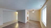 This single-floor space features an open layout that seamlessly connects the main living area to the kitchen. The living room is finished with durable wood-style flooring in a rich, dark tone, complemented by neutral-painted walls and a textured popcorn ceiling. A ceiling fan with a multi-bulb light fixture is centrally mounted, providing both air circulation and ambient lighting throughout the space.

A partial dividing wall separates the living area into distinct zones, with one section transitioning to lighter tile-style flooring, offering a natural delineation between areas. White baseboards run consistently throughout, adding a clean, finished look.

The adjacent kitchen is accessible via an open passthrough and features warm wood-toned cabinetry, a white range, and light-colored countertops. A doorway visible from the main living area leads to additional rooms beyond, indicating a functional and connected floor plan. The overall ambiance is clean, neutral, and move-in ready, with simple finishes that provide a versatile backdrop for a variety of interior styles.