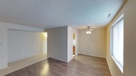 This single-floor space features an open layout that seamlessly connects the main living area to the kitchen. The living room is finished with durable wood-style flooring in a rich, dark tone, complemented by neutral-painted walls and a textured popcorn ceiling. A ceiling fan with a multi-bulb light fixture is centrally mounted, providing both air circulation and ambient lighting throughout the space.

A partial dividing wall separates the living area into distinct zones, with one section transitioning to lighter tile-style flooring, offering a natural delineation between areas. White baseboards run consistently throughout, adding a clean, finished look.

The adjacent kitchen is accessible via an open passthrough and features warm wood-toned cabinetry, a white range, and light-colored countertops. A doorway visible from the main living area leads to additional rooms beyond, indicating a functional and connected floor plan. The overall ambiance is clean, neutral, and move-in ready, with simple finishes that provide a versatile backdrop for a variety of interior styles.