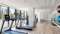 The fitness room at 2060 Quingate Place in Halifax, Nova Scotia, is a well-appointed exercise facility designed to support a comprehensive workout experience. The space is thoughtfully laid out across a single floor, offering generous room for a wide variety of equipment and activities.

Floor-to-ceiling windows line the perimeter, flooding the interior with abundant natural light and providing open views of the surrounding greenery and streetscape. The light hardwood-style flooring complements the clean, white walls and recessed drop-tile ceiling, creating a bright and modern aesthetic throughout.

The facility is equipped with multiple treadmills, elliptical trainers, and a cable machine from Hoist, catering to both cardio and functional strength training. A dedicated free weights area features a full dumbbell rack, while additional strength machines and benches support a range of resistance exercises. Large wall-to-wall mirrors on one side of the room enhance the sense of openness and allow users to monitor their form during workouts.

The space also accommodates flexibility and balance training, with a stability ball visible in one corner. The overall layout is open and uncluttered, ensuring ease of movement between equipment. The combination of ample natural light, quality equipment, and a clean, contemporary design makes this fitness room a functional and inviting environment for residents.