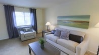 This two-floor residence offers a thoughtfully designed living space with a calm, contemporary aesthetic. The living room serves as the heart of the main level, finished in soft neutral tones that create a bright and inviting atmosphere. A large window dressed with dark grayish-blue curtain panels allows natural light to fill the room, complementing the light-colored walls throughout. The space accommodates a generously sized sofa adorned with patterned accent pillows in navy and gray tones, paired with a matching armchair, a dark wood coffee table, and a side table with a warm table lamp. A large landscape-inspired artwork anchors one wall, adding a subtle coastal touch to the overall decor. The carpeted flooring adds warmth underfoot, while the open layout allows for comfortable furniture arrangement and easy movement throughout the room. The two-floor layout suggests additional living or sleeping quarters on the upper level, offering separation between common and private areas. Overall, the space presents a clean, understated design with a cohesive color palette that balances comfort and style.