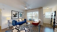 This single-floor residence is designed around an open-concept layout that seamlessly connects the living, dining, and kitchen areas, creating a cohesive and airy atmosphere throughout. Light wood-tone flooring flows consistently across the main living spaces, complementing the crisp white walls and modern finishes.

The living room is a well-lit, comfortable space anchored by large windows that invite natural light and offer outdoor views. A ceiling fan overhead and bold artwork on the walls add character, while the generous floor area accommodates a full furniture arrangement with ease.

The dining area sits adjacent to the living room, bathed in natural light from a wall of windows. It offers a dedicated space for entertaining, with room for a full dining set and sideboard storage.

The kitchen features warm wood cabinetry, stainless steel appliances including a refrigerator, microwave, and range, and light countertops. A kitchen island with bar seating serves as a functional and social centerpiece connecting the kitchen to the main living area.

The bedroom is a calm, carpeted retreat with dual windows, ample natural light, and room for substantial furnishings. The bathroom includes a full soaking tub with marble-style tile surround, maintaining a clean and contemporary aesthetic throughout.
