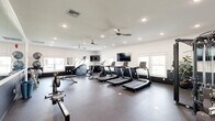 Located at 100 Bill Hegeman Blvd in Conway, Arkansas, this fitness center offers a comprehensive workout environment within a single, open-plan space. The room is finished with a durable rubber flooring surface suitable for high-traffic exercise use, and the crisp white walls are accented by a dark wainscoting detail that gives the space a clean, modern aesthetic.

Natural light flows in through multiple windows positioned along the walls, complementing the recessed ceiling lighting and dual ceiling fans that help maintain air circulation throughout the room. A large flat-screen television is mounted on the wall, providing entertainment during workouts.

The cardio zone is well-appointed with a row of treadmills, an elliptical trainer, a stair climber, and a stationary cycle. For strength training, the space includes a multi-functional cable machine, a rack of dumbbells spanning a wide weight range, a set of medicine/stability balls, and an adjustable weight bench. Two rowing machines round out the equipment selection, catering to full-body conditioning workouts.

A full-length wall mirror along one side of the room visually expands the space and allows users to monitor their form. The overall layout provides ample open floor space between equipment stations, supporting a comfortable and functional fitness experience.