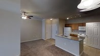 This single-story residence in Lewisville, TX offers an open-concept floor plan that integrates the living area and kitchen into one cohesive space. The living area is finished with wall-to-wall carpet, crown molding, and a ceiling fan with a built-in light fixture, contributing to a comfortable and functional atmosphere. Neutral-toned walls complement the understated design throughout.

The kitchen is positioned along one side of the open layout and features a peninsula counter with a granite-style surface, providing both workspace and a casual dining or serving area. Medium-toned wood cabinetry offers ample storage, while a white refrigerator and matching appliances complete the setup. Track lighting above the kitchen area ensures focused illumination for meal preparation.

A paneled entry door is visible from the main living space, accompanied by what appears to be a closet nearby, adding practical storage to the layout. The overall design is clean and straightforward, with consistent finishes throughout, making this a functional and well-organized single-floor living space.