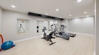 Located at 343 W Fourth St in Williamsport, Pennsylvania, this dedicated fitness room offers a clean, functional space designed for cardiovascular exercise and general wellness. The room features a neutral gray and white color palette with smooth, light-toned hardwood-style flooring that contributes to a bright, modern atmosphere. Recessed ceiling lighting provides consistent, well-distributed illumination throughout the space. A large wall of mirrors along one side enhances the sense of openness and allows users to monitor their form during workouts. The room is equipped with a selection of commercial-grade cardio machines, including a treadmill, elliptical trainer, stationary bike, and stair-stepper, all from reputable fitness brands. An exposed HVAC duct runs along the ceiling, adding an industrial accent to the otherwise clean-lined interior. The layout provides ample open floor space beyond the equipment area, allowing for stretching or floor exercises. A dedicated exit door with emergency signage ensures safety compliance. Structural columns are neatly integrated into the design without disrupting the usable floor area. This well-appointed fitness room serves as a convenient, fully self-contained exercise facility.