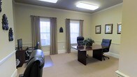 This single-floor office space is designed to accommodate professional work in a refined, well-appointed environment. The room is finished with soft yellow-green walls, crisp white crown molding, and a chair rail that adds a classic architectural detail. Neutral carpet flooring runs throughout, contributing to a quiet, comfortable atmosphere.

At the center of the space sits a substantial dark wood executive desk accompanied by black leather office chairs, creating a distinguished focal point. A decorative plant atop the desk adds a touch of warmth and life to the setting. Two large windows dressed with beige drape panels and white horizontal blinds allow generous natural light to fill the room while maintaining privacy. Framed artwork on the walls completes the polished, professional aesthetic.

Overhead, a flush-mounted rectangular light fixture provides consistent ambient illumination, complementing the natural light from the windows. The overall design balances functionality with formal elegance, making it well-suited for executive meetings, client consultations, or focused individual work.