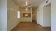 This single-floor space offers a straightforward and functional layout designed for comfortable everyday living. The main living area is finished with warm wood-look flooring that extends throughout, creating a cohesive and inviting atmosphere. A ceiling fan with an integrated light fixture is centrally positioned, providing both air circulation and ambient lighting. Natural light enters through a vertical-blind-covered window near the entryway, brightening the open living space.

The kitchen is accessible via a pass-through counter that connects it to the living area, making the space feel open and communicative. It features warm wood-toned upper and lower cabinetry, a range with overhead ventilation, and a full-size refrigerator. This setup provides a practical cooking and dining environment.

A hallway visible from the main living area leads further into the unit, suggesting additional private spaces beyond the main room. Neutral, light-colored walls throughout contribute to a clean and airy feel, while white trim and doors offer a crisp contrast. The overall design is practical and unpretentious, making this a functional space well-suited for daily living.