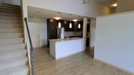 This two-storey space showcases a well-appointed kitchen as its central feature. The kitchen is designed with rich, dark espresso-toned cabinetry that extends to the upper walls, providing ample storage. A central island with a light stone countertop serves as both a functional workspace and a casual gathering point, complete with an integrated sink. Pendant lighting hangs above the island, adding focused illumination and a contemporary design touch, while recessed lighting throughout the kitchen ensures the space remains bright and practical.

Stainless steel appliances, including a full-sized refrigerator, are seamlessly integrated into the cabinetry, contributing to the sleek, modern aesthetic. The kitchen transitions naturally into the surrounding open-plan area, which features light hardwood-style flooring that runs consistently throughout, creating a sense of continuity and openness. A structural column near the kitchen adds an architectural element to the layout.

The space benefits from an open connection between its two floors, with staircase railings visible, hinting at the vertical flow of the layout. Neutral wall tones complement the contrast between the dark cabinetry and light flooring, resulting in a balanced and inviting overall ambiance.