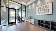 Located at 3030 Ascend Oak St in Apopka, Florida, this well-appointed fitness center spans a generous single-floor layout designed to accommodate a comprehensive workout experience. The main exercise area features high tray ceilings with recessed lighting and large black-framed windows that flood the space with natural light and offer views of the surrounding greenery. The floor is finished with durable, patterned carpet tiles that define the open workout zones. A full complement of strength and cardio equipment fills the space, including cable machines, a Smith machine, adjustable benches, dumbbell racks, and a variety of selectorized machines. A wall-mounted television is positioned for visibility throughout the floor. A dedicated cycling studio is enclosed by dramatic floor-to-ceiling black-framed glass walls and a glass door, creating a visually open yet acoustically separate space. Inside, stationary cycling bikes are arranged around a vibrant, large-scale abstract wall mural that energizes the room. The studio is also equipped with a ceiling fan. Adjacent to the cycling studio entrance, a hydration station with a water fountain and under-counter refrigerator is built into a sleek gray cabinetry unit, complemented by curated wall art. The overall aesthetic is clean and contemporary, with a soft blue and gray color palette throughout.