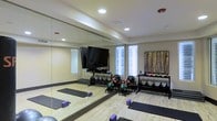 This dedicated fitness studio is thoughtfully designed to support a wide range of workout routines. The space features light hardwood flooring that lends warmth and a clean aesthetic, while a full-length mirrored wall visually expands the room and allows for form monitoring during exercise. Recessed ceiling lighting provides bright, even illumination throughout the space. A large flat-screen TV is mounted on the mirrored wall, ideal for streaming workout classes or training videos. Natural light filters in through multiple windows fitted with shades, enhancing the open and energizing atmosphere. The studio is well-stocked with a comprehensive selection of equipment, including a tiered dumbbell and kettlebell rack with varying weights, a collection of medicine balls stored on dedicated racks, and yoga mats and foam blocks arranged on the floor for stretching and flexibility work. A decorative wall-mounted art piece adds a touch of character to the space. The room is served by a ceiling-mounted HVAC system and includes a fire suppression sprinkler for safety. Overall, this fitness studio offers a functional, organized, and motivating environment suitable for strength training, cardio, and mind-body workouts alike.