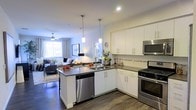 This single-floor residence showcases a thoughtfully designed open-concept layout that seamlessly connects the kitchen and living areas into one cohesive, inviting space. The kitchen is a standout feature, outfitted with crisp white cabinetry, dark countertops, and a full suite of stainless steel appliances, including a gas range, built-in microwave, and dishwasher. A peninsula island with an integrated sink serves as both a functional workspace and a casual dining spot, illuminated overhead by two elegant pendant lights. A decorative tile backsplash adds a refined touch to the kitchen's clean aesthetic. The flooring throughout is rich, dark hardwood, providing a striking contrast to the white cabinetry and lending warmth to the space. The adjoining living area is generously proportioned and bathed in natural light from large windows dressed with neutral drapes. A ceiling fan adds comfort to the room, which accommodates a full seating arrangement with ease. The overall ambiance is modern and welcoming, with a neutral palette, recessed lighting, and thoughtful design details that create a polished yet comfortable everyday living environment.