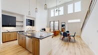 This contemporary two-story residence in Dallas, TX offers a striking open-concept design anchored by dramatic double-height ceilings in the main living area. Light wood flooring runs throughout, enhancing the bright, airy atmosphere created by an abundance of natural light streaming through tall windows. The kitchen is a standout feature, equipped with bold navy and natural wood cabinetry, dark countertops, stainless steel appliances, a built-in wine cooler, and a large center island with bar seating — ideal for both casual dining and entertaining. The kitchen flows seamlessly into the dining and living areas, where soaring ceilings and a ceiling fan add to the sense of volume and comfort. The spacious primary bedroom is located on the upper level and features an open interior window overlooking the double-height living space below, bringing in additional light and architectural interest. It also includes ample closet space. The second bedroom is equally well-appointed with its own walk-in closet. The bathroom offers a combination tub and shower with decorative tile surround, while a secondary bathroom features dark cabinetry and generous storage. A hallway connects the upstairs rooms, completing this well-designed and modern Dallas home.