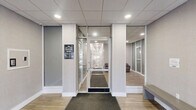 Located at 570 W Dekalb Pike in King of Prussia, PA, this single-floor commercial space offers a polished and professional environment designed to make a strong first impression. The entrance features large glass panels flanking a central glass door marked with the building's address, flooding the interior with natural light and offering clear sightlines into the space beyond.

Inside, the lobby transitions seamlessly into a well-appointed hallway, both finished with light wood-look flooring that lends warmth to the otherwise neutral palette of soft gray walls and white trim. Dark charcoal entry mats provide a practical yet refined contrast at key thresholds. A standout feature of the lobby area is the cross-shaped contemporary light fixture mounted to the drop-tile ceiling, adding a modern design element to the space.

A comfortable waiting area is furnished with deep burgundy leather armchairs and a glass side table, positioned alongside framed black-and-white artwork — conveying a sophisticated, professional atmosphere. Wall-mounted sconce lighting accents the seating area with a warm, welcoming glow. Frosted and clear glass partitions throughout create visual separation between zones while maintaining an open, airy feel. A decorative boxwood plant in a dark planter adds a touch of greenery, completing the clean, contemporary aesthetic of this well-maintained commercial suite.