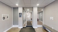 Located at 570 W Dekalb Pike in King of Prussia, PA, this single-floor commercial space offers a polished and professional environment designed to make a strong first impression. The entrance features large glass panels flanking a central glass door marked with the building's address, flooding the interior with natural light and offering clear sightlines into the space beyond.

Inside, the lobby transitions seamlessly into a well-appointed hallway, both finished with light wood-look flooring that lends warmth to the otherwise neutral palette of soft gray walls and white trim. Dark charcoal entry mats provide a practical yet refined contrast at key thresholds. A standout feature of the lobby area is the cross-shaped contemporary light fixture mounted to the drop-tile ceiling, adding a modern design element to the space.

A comfortable waiting area is furnished with deep burgundy leather armchairs and a glass side table, positioned alongside framed black-and-white artwork — conveying a sophisticated, professional atmosphere. Wall-mounted sconce lighting accents the seating area with a warm, welcoming glow. Frosted and clear glass partitions throughout create visual separation between zones while maintaining an open, airy feel. A decorative boxwood plant in a dark planter adds a touch of greenery, completing the clean, contemporary aesthetic of this well-maintained commercial suite.