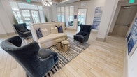 This thoughtfully designed single-floor community space combines coastal elegance with modern functionality. The main lounge area sets the tone with a sophisticated coffered ceiling accented in teal and gold trim, a striking sputnik chandelier, and light wood-look tile flooring that flows throughout. Plush navy velvet accent chairs and a cream sofa arranged around metallic accent tables create a welcoming gathering spot, complemented by large-scale ocean-themed artwork that reinforces the coastal aesthetic.

Adjacent to the lounge, a bright dining area features a round glass-top table surrounded by teal upholstered chairs, topped by a bold geometric gold chandelier. Expansive windows flood the space with natural light and offer views of the surrounding outdoor areas.

The kitchen and entertaining bar is well-appointed with granite countertops, white cabinetry, stainless steel appliances including a beverage refrigerator and dishwasher, and a large island with bar seating. A textured wave-pattern backsplash adds visual interest, while a wall-mounted television enhances the space for social gatherings.

A dedicated workspace is built into the kitchen area, featuring a built-in desk with dual monitors, task lighting, and bar-height seating — ideal for productivity. Throughout, the design maintains a cohesive coastal palette of navy, cream, and gold, creating a polished and inviting atmosphere.