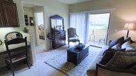 This single-floor residence showcases a thoughtfully designed living space with a classic, refined aesthetic. The living room serves as the heart of the home, anchored by a dark wood storage trunk used as a coffee table and complemented by an ornate, carved wooden dresser topped with a decorative mirror, candleholders, and a globe accent piece. A cushioned accent chair in a neutral tone adds to the traditional charm of the room. Plush carpeting and a patterned area rug contribute warmth underfoot, while large sliding glass doors flood the space with abundant natural light. These doors open to a private balcony furnished with a bistro-style table and chairs, offering views of surrounding greenery and a neighboring building. The open layout allows a clear sightline into an adjoining bedroom, which features similarly soft, neutral tones and carpeted flooring, maintaining a cohesive design throughout. The overall ambiance is warm and inviting, blending traditional furnishings with comfortable, livable spaces that feel both elegant and functional.
