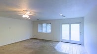 This single-floor space offers a clean, open layout designed for comfortable everyday living. The main living area is finished with wall-to-wall carpeting that transitions to tile flooring near the French doors, creating a practical and visually distinct entryway to the outdoors. The French doors serve as a prominent architectural feature, flooding the room with natural light while providing direct access to the exterior. A standard window with horizontal blinds on the adjacent wall adds an additional source of daylight, contributing to the room's bright and airy atmosphere. The ceiling is fitted with a multi-blade ceiling fan with an integrated light fixture, offering both ventilation and overhead illumination. A recessed light is also installed, supplementing the lighting scheme with a clean, modern touch. Walls are finished in a neutral white tone, providing a versatile backdrop suitable for a variety of interior styles. Visible HVAC vents indicate a central air system throughout. The overall ambiance is light, functional, and well-suited for a range of living arrangements.