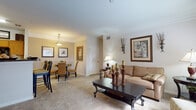 Located in Wesley Chapel, Florida, this well-designed single-floor residence offers a thoughtfully arranged layout with a warm, inviting ambiance throughout. The living room serves as the heart of the home, featuring plush carpeting, crown molding, and large windows that flood the space with natural light while framing views of the surrounding greenery. The open flow between the living and dining areas creates a seamless entertaining space, with the dining room accommodating a round glass-top table beneath a classic ceiling fixture, accented by tasteful framed artwork on neutral-toned walls. The galley-style kitchen is efficiently designed with warm wood cabinetry, dark granite-style countertops, tile flooring, and black appliances including a refrigerator, range, microwave, and dishwasher. Both bedrooms are generously proportioned with soft carpeting, ample closet space, and windows that invite natural light. The primary bathroom features an extended vanity with dark granite countertops, white cabinetry, and a large mirror, while the secondary bathroom includes a full tub-shower combination with clean white tile surround. Together, these spaces deliver a cohesive and comfortable living environment suited for everyday life in Wesley Chapel.