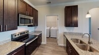 This single-floor residence offers a thoughtfully designed layout that blends functionality with modern finishes throughout. The kitchen is a standout feature, showcasing rich dark-espresso cabinetry, light granite countertops, and a full suite of stainless steel appliances including a refrigerator, electric range, and over-the-range microwave. A peninsula with pendant lighting above serves as both a prep space and casual dining area, opening directly into the living space. The living area is generously sized, finished with neutral-toned carpet and crown molding that adds a refined touch to the overall aesthetic. A ceiling fan and multiple windows allow for natural light and comfortable airflow. Adjacent to the kitchen, a dedicated laundry room houses a full-size side-by-side washer and dryer, adding everyday convenience. The bedroom is a calm, well-proportioned retreat with plush carpet, a window with exterior views, and a textured ceiling. The bathroom is finished with the same cohesive design language — dark cabinetry, granite countertop vanity, a large mirror with overhead lighting, and a tub-shower combination featuring marble-style tile surrounds. Light wood-look flooring in the kitchen and bath areas ties the space together seamlessly.