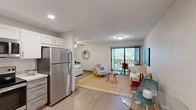 This thoughtfully designed studio in Port Charlotte, Florida, offers a smart, efficient floor plan that maximizes every square foot. The open-concept living area seamlessly connects the bedroom, living, and dining spaces, creating a bright and airy atmosphere enhanced by light wood-style flooring and a ceiling fan overhead. Sliding glass doors lead to a private balcony, flooding the space with natural light and offering outdoor views.

The kitchen is a standout feature, boasting white shaker upper cabinets, contrasting grey lower cabinets, light granite-style countertops, a subway tile backsplash, and a full suite of stainless steel appliances including a refrigerator, range, microwave, and dishwasher. A dedicated dining nook near the entrance accommodates a glass-top table and seating.

The spacious bedroom area comfortably fits a full bed with built-in shelving and benefits from a ceiling fan and ample room to move. A dedicated closet provides convenient storage. The two bathrooms are clean and modern, one featuring a walk-in shower with brushed gold fixtures, and the other a vanity with a white quartz countertop, grey cabinetry, and a large mirror. The overall aesthetic carries a light coastal charm throughout.
