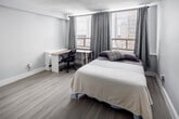 Located on the 9th floor at 186 King Street in downtown London, Ontario, this one-bedroom, one-bathroom unit offers a well-organized layout that maximizes every square foot of its compact footprint. Light wood-tone laminate flooring runs throughout, lending a clean, consistent aesthetic across the entire space.

The open-area kitchen is positioned near the entrance and features white shaker-style cabinetry, a white subway tile backsplash, a stainless steel refrigerator, a gas range, and an under-cabinet range hood. A single-basin sink and modest counter space complete the functional cooking area. The adjacent electrical panel is neatly integrated into the entrance corridor.

The bedroom is the most generously sized room in the unit, accommodating a full bed with space remaining for a desk and additional furnishings. Two large windows allow notable natural light and frame views of the surrounding downtown London cityscape, with grey curtains providing privacy and light control.

The bathroom features a white vanity with drawers, a toilet, and a shower-tub combination with a curtain, all set on matching light wood-tone flooring with crisp white walls. The overall aesthetic throughout the unit is neutral, modern, and functional, suited for single occupancy in an urban setting.