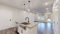 This single-floor unit presents a clean, contemporary design highlighted by wood-look plank flooring that flows consistently throughout the entire space. The open-concept kitchen serves as the centerpiece, anchored by a large island with quartz countertops, an undermount stainless steel farmhouse sink, and a matte black pull-down faucet. Pendant lighting with glass fixtures hangs above the island, adding a refined touch. White shaker-style cabinetry with black hardware lines the kitchen walls, complemented by a classic subway tile backsplash and a full suite of stainless steel appliances, including a refrigerator, range, microwave, and dishwasher. The kitchen opens directly into a generously sized living area bathed in natural light from large windows fitted with roller shades, with a door providing access to an outdoor balcony. The bedrooms are well-proportioned, each featuring neutral tones, recessed lighting, and windows with exterior views. The primary bathroom is notably spacious, offering an elongated dual-sink vanity with quartz countertops, matte black fixtures, an illuminated mirror, a glass-door walk-in shower, and a walk-in closet accessible directly from the bathroom. The overall aesthetic is modern and cohesive throughout.