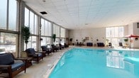This indoor pool facility offers a well-appointed aquatic environment designed for both leisure and relaxation. The centerpiece is a generously sized swimming pool with clear, blue water, surrounded by a wide tiled deck that provides ample circulation space. Along one wall, a sweeping row of floor-to-ceiling windows floods the area with natural light, offering views of the exterior while creating an open, airy atmosphere despite being fully enclosed.

The high drop-panel ceiling adds to the sense of vertical space, while the white-painted brick walls on the opposite side lend a clean, classic aesthetic to the interior. A dedicated lounge area lines the perimeter of the pool, furnished with wicker and upholstered seating arrangements, potted plants, and small accent tables, encouraging relaxation before or after a swim. The combination of natural light, greenery, and comfortable furnishings gives the space a welcoming, resort-like character. Overall, the space balances functional aquatic amenities with a comfortable social environment suitable for year-round use.