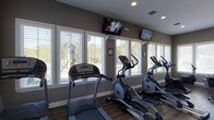 This single-floor fitness center is thoughtfully designed to provide a bright and motivating workout environment. The space is lined with large windows dressed with white horizontal blinds, allowing abundant natural light to fill the room while offering pleasant views of the surrounding greenery outdoors. The flooring features rich, dark hardwood-style planks that add a polished, modern touch to the interior.

The cardio area is well-appointed with multiple treadmills and elliptical machines arranged in a row, making efficient use of the available floor space. Two wall-mounted flat-screen televisions are positioned at optimal viewing angles, ensuring that users can stay entertained or informed during their workouts. Recessed lighting in the ceiling supplements the natural light, keeping the space well-lit at all times.

The neutral gray walls contribute to a clean and focused atmosphere, while the white trim and window frames create a crisp contrast throughout the room. Ventilation is supported by ceiling vents, ensuring a comfortable environment during exercise. The overall ambiance is practical yet inviting, offering a functional and well-maintained setting suited for daily fitness routines.