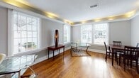 Located at 1421 Roper Mountain Rd in Greenville, South Carolina, this single-story home offers a thoughtfully designed interior with a cohesive blend of classic and contemporary details throughout. Rich hardwood flooring flows seamlessly across the main living areas, anchoring the warm, welcoming atmosphere of the space.

The dining area features crisp white crown molding and neutral gray walls, creating an elegant yet understated setting. A doorway connects it to adjacent rooms, including a space with tile flooring and a staircase detail visible in the background, adding architectural interest to the layout.

A standout feature of the home is a spacious, light-filled room that functions as a home office or flex space. This room is distinguished by a distinctive tray ceiling fitted with built-in ambient uplighting that casts a warm golden glow, complementing the gleaming hardwood floors below. Large windows with white plantation shutters line multiple walls, inviting generous natural light and offering pleasant views of the surrounding greenery and neighborhood.

The same room opens toward a well-appointed kitchen area with white cabinetry visible through a connecting doorway, suggesting a practical and accessible floor plan. The neutral gray palette, clean white trim work, and thoughtful lighting design give the entire home a polished, move-in-ready appeal suitable for a variety of lifestyles.