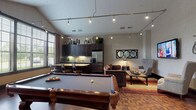Located at 3280 Mill Run Dr in Hilliard, Ohio, this single-floor community clubroom offers a well-appointed gathering space designed for socializing and recreation. The open-concept layout seamlessly integrates multiple functional zones, creating a versatile environment for residents.

At the heart of the space is a full-size billiards table set atop rich, parquet-style hardwood flooring that extends throughout the room. Industrial-style track lighting runs along the ceiling, casting warm Edison-bulb light across the entire area and contributing to the space's modern yet inviting ambiance.

The fully equipped kitchen features dark espresso cabinetry, granite countertops, a stainless steel refrigerator, dishwasher, microwave, and a double-basin sink. A kitchen island with bar seating provides additional gathering space. Large windows with slatted blinds allow natural light to filter in, offering views of the surrounding greenery.

The lounge area is thoughtfully arranged with plush brown sofas, upholstered wingback chairs, and a rustic wooden coffee table set atop a warm-toned area rug. A wall-mounted flat-screen television and decorative wall art — including woven circular accents and a metal bicycle sculpture — add character. A framed triple-circle mirror completes the sophisticated, layered aesthetic of this welcoming community retreat.