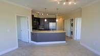 This single-floor residence offers a well-designed open-concept layout that seamlessly connects the kitchen and living areas, creating a spacious and functional environment. The kitchen is a standout feature, anchored by a generously sized curved island with a light countertop and an integrated sink, providing ample workspace and serving as a natural focal point for the room. Dark espresso cabinetry lines the kitchen walls, offering abundant storage and a rich contrast to the lighter countertops and neutral-toned walls. Stainless steel appliances, including a refrigerator, stove, and microwave, add a sleek, contemporary finish. Track lighting runs across the ceiling, complementing stylish pendant lights that hang above the island to create warm, layered illumination throughout the space. Crown molding along the ceiling adds a refined architectural touch. The adjacent living area features soft carpeting underfoot, contributing to a comfortable and inviting atmosphere. A dedicated in-unit laundry closet with a stacked washer and dryer provides convenient, practical utility. The overall design balances modern aesthetics with everyday functionality, making this a thoughtfully appointed living space.