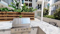 This beautifully designed outdoor courtyard serves as a premier amenity space nestled within a contemporary multi-story residential building. The space is thoughtfully laid out with clean, light-toned concrete pathways and open-air gathering areas that invite residents to relax and socialize in a well-maintained outdoor environment.

At the heart of the courtyard is a fully equipped outdoor kitchen, anchored by a professional-grade Viking stainless steel built-in grill and complemented by elegant granite countertops with distinctive veining. A warm wood-slatted privacy screen backs the kitchen station, adding a natural, textural contrast to the otherwise modern aesthetic. Adjacent bar-height seating and a pergola structure provide a shaded lounge area, ideal for casual dining and entertaining.

The courtyard is further enhanced by an abundance of thoughtfully curated landscaping, including raised concrete planters filled with mature trees, tropical plants, and seasonal greenery. These verdant elements soften the architecture and create a sense of privacy and tranquility throughout the space.

Flanked on multiple sides by the building's balconied upper floors, the courtyard benefits from an open-sky orientation that allows generous natural light to flood the area throughout the day. Low-profile step lighting integrated into the planter walls adds a refined finishing touch to the overall design.