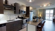 This contemporary single-floor space showcases a thoughtfully designed open-concept layout that seamlessly connects the kitchen and living areas. The kitchen is a standout feature, outfitted with dark espresso cabinetry that extends to the ceiling for generous storage, complemented by crisp white countertops and a stylish tile backsplash in a neutral mosaic pattern. A full suite of stainless steel appliances — including a French door refrigerator, electric range with microwave above, and an undermount sink — provides a sleek, modern aesthetic. A peninsula with a contrasting countertop and bar seating extends the kitchen into the living space, making it ideal for dining and entertaining. The open living area, visible from the kitchen, is bright and welcoming, illuminated by ceiling-mounted lighting and a warm floor lamp that creates an inviting ambiance. Light wood-toned flooring runs throughout, tying the spaces together with a cohesive, contemporary feel. The overall design balances functionality and style, with clean lines, neutral tones, and high-quality finishes that lend the space a polished, modern character.