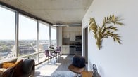 This convertible unit is thoughtfully designed to maximize both functionality and style within a single-floor layout. The space is immediately defined by its dramatic floor-to-ceiling windows, which flood the interior with abundant natural light and frame expansive, high-rise views of the surrounding cityscape — a standout architectural feature that anchors the entire living area.

The open-concept layout seamlessly connects the living, dining, and kitchen zones. The kitchen is equipped with stainless steel appliances, including a range and microwave, complemented by light wood cabinetry and ample counter space. It flows naturally into a compact dining area positioned directly beside the oversized windows, making everyday meals feel elevated by the panoramic backdrop.

Exposed concrete ceilings add an industrial character to the space, contrasting with the clean white walls and warm wood-tone flooring that run throughout. Decorative touches, such as the sculptural gold botanical wall piece, lend an artistic sensibility to the interior. The living area accommodates seating comfortably, completing a well-rounded, versatile layout suited to urban living.