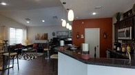 This single-floor space showcases a thoughtfully designed open-concept layout that seamlessly connects the kitchen, dining, and living areas. The kitchen features a curved bar-style counter finished with dark granite-look surfaces, offering casual seating and a natural gathering point for the space. A trio of modern pendant lights hang above the counter, adding warm, focused illumination and a contemporary design element. The kitchen flows effortlessly into the dining area, which includes a bistro-style table with bar stools, perfect for everyday dining. A striking terracotta-orange accent wall anchors the space, adding depth and warmth to the otherwise neutral palette of whites and grays. Recessed lighting throughout the ceiling ensures consistent brightness across the open floor plan. The living area is visible beyond the dining space and features a comfortable seating arrangement with a sofa, accent chairs, and a flat-screen television, all grounded by a bold geometric-patterned rug over rich hardwood-style flooring. Wall art and decorative accents throughout reflect an urban, cosmopolitan aesthetic, giving the space a curated and personalized character. Overall, the layout is efficient, stylish, and well-suited for both everyday living and casual entertaining.