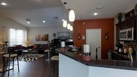 This single-floor space showcases a thoughtfully designed open-concept layout that seamlessly connects the kitchen, dining, and living areas. The kitchen features a curved bar-style counter finished with dark granite-look surfaces, offering casual seating and a natural gathering point for the space. A trio of modern pendant lights hang above the counter, adding warm, focused illumination and a contemporary design element. The kitchen flows effortlessly into the dining area, which includes a bistro-style table with bar stools, perfect for everyday dining. A striking terracotta-orange accent wall anchors the space, adding depth and warmth to the otherwise neutral palette of whites and grays. Recessed lighting throughout the ceiling ensures consistent brightness across the open floor plan. The living area is visible beyond the dining space and features a comfortable seating arrangement with a sofa, accent chairs, and a flat-screen television, all grounded by a bold geometric-patterned rug over rich hardwood-style flooring. Wall art and decorative accents throughout reflect an urban, cosmopolitan aesthetic, giving the space a curated and personalized character. Overall, the layout is efficient, stylish, and well-suited for both everyday living and casual entertaining.
