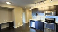 This single-floor space showcases a thoughtfully designed open-concept layout that seamlessly connects the kitchen and living areas. The kitchen is a standout feature, outfitted with sleek dark cabinetry that contrasts beautifully against a light marble-style tile backsplash and crisp white countertops. Stainless steel appliances, including a dishwasher and range, lend a polished, modern aesthetic, while a deep undermount sink adds both style and function. Track lighting mounted on the ceiling provides targeted illumination throughout the kitchen, highlighting the contemporary design details. Rich, dark hardwood flooring flows throughout the space, adding warmth and cohesion to the open floor plan. The adjoining living area is bright and airy, featuring neutral-toned walls that create a versatile backdrop for a variety of interior styles. A hallway leads to additional rooms, with clean white-paneled doors and functional storage solutions visible in the background. A floor-level vent ensures comfortable climate control throughout. The overall ambiance is modern and refined, combining practical design with upscale finishes to create a welcoming and well-appointed living environment.