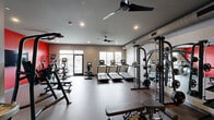 Located at 9 Presidential Blvd in Bala Cynwyd, PA, this fitness center offers a thoughtfully designed exercise space that accommodates a comprehensive range of workout routines. The room features durable rubber flooring throughout, providing both safety and comfort during exercise. Ceiling fans and recessed lighting contribute to a well-ventilated and well-lit environment, while multiple windows along the perimeter allow natural light to fill the space and offer views of the surrounding area.

The cardio zone is prominently equipped with a row of Matrix treadmills featuring integrated display screens, complemented by a recumbent bike and additional cardio machines positioned to maximize floor space. A dedicated strength training area houses a multi-functional cable machine set against a bold red accent wall, which serves as a visual focal point and energizes the room's overall aesthetic. A full dumbbell rack with a wide weight range lines one wall, paired with floor-length mirrors that enhance the sense of openness and allow users to monitor their form.

Additional equipment includes a rowing machine, a weight bench, and a wall-mounted flat-screen television. A water fountain is conveniently integrated into the space. The neutral gray and white walls, accented by the striking red feature wall, create a motivating and modern atmosphere suited for a variety of fitness levels and training styles.