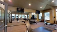This fitness center offers a comprehensive workout environment across a single-story layout. The space is outfitted with a diverse selection of exercise equipment, including cardio machines such as treadmills, ellipticals, and a stationary bike, as well as a cable/weight machine for strength training. A full dumbbell rack with a range of weights and an adjustable workout bench provide additional options for free-weight training. Medicine balls and stability balls are neatly organized along one wall, catering to functional fitness routines. Large mirrors line the walls, enhancing the sense of space while allowing users to monitor their form. Two wall-mounted flat-screen televisions are positioned for easy viewing from multiple areas of the room. Recessed lighting keeps the space well-lit, complemented by natural light streaming through a series of windows along the perimeter, offering views of surrounding greenery. The carpeted flooring and warm, neutral-toned walls contribute to a comfortable and functional atmosphere suited for a wide range of fitness activities.