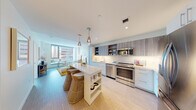 This single-floor residence offers a thoughtfully designed open-concept layout that seamlessly connects the kitchen, dining, and living areas. The kitchen is a standout feature, showcasing two-tone cabinetry with wood-grain upper cabinets and clean white lower cabinets, a white subway tile backsplash, and a full suite of stainless steel appliances including a French door refrigerator, range, dishwasher, and built-in microwave. A white island with integrated shelving and woven bar stools serves as both a functional workspace and casual dining spot, illuminated by elegant pendant lights overhead.

The living area is bright and airy, benefiting from expansive floor-to-ceiling windows that frame dynamic urban cityscape views. Light wood-tone flooring flows consistently throughout the space, enhancing the clean, contemporary aesthetic.

Two well-proportioned bedrooms each feature large windows with city views, allowing abundant natural light. The primary bedroom connects to a sleek en-suite bathroom appointed with white cabinetry, a backlit vanity mirror, and quartz countertops. The second bedroom is equally bright and well-appointed. The overall ambiance is modern, minimalist, and urbane, with a neutral palette accented by warm tones throughout.