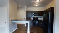 This single-floor space is designed with an open-concept layout that integrates the kitchen and living areas into one cohesive environment. The kitchen is a standout feature, outfitted with rich, dark espresso-toned cabinetry that extends from floor to ceiling, providing ample storage. Black appliances, including a gas range, over-the-range microwave, and refrigerator, complement the cabinetry and contribute to a consistent, modern aesthetic. A decorative tile backsplash adds a subtle textural detail along the cooking wall.

A white peninsula counter with a gray laminate top serves as the central dividing element between the kitchen and the adjoining living space. Its clean white base contrasts effectively with the darker kitchen elements and provides a functional surface for dining or food preparation. Dark hardwood-style flooring runs throughout, anchoring the space with warmth and continuity. The walls are finished in neutral tones, and recessed and flush-mount ceiling lighting ensures the interior remains well-lit. The layout offers a practical, connected flow between living and cooking zones.