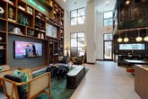 This contemporary communal lounge is defined by its dramatic double-height ceilings and an open, airy layout that fosters both relaxation and social interaction. Large black-framed windows flood the space with natural light while offering views of the surrounding outdoor area. A floor-to-ceiling warm wood bookshelf wall anchors one side of the room, displaying curated décor, artwork, and a wall-mounted flat-screen TV, accented by a glowing neon "Good Vibes Only" sign that adds a playful, modern touch. Comfortable lounge seating is thoughtfully arranged on a rich green area rug, featuring a mix of upholstered chairs, a sleek leather bench, and eclectic accent furniture. A striking industrial-style communal table with wooden swivel stools provides a versatile workspace or gathering area beneath an artistic wire chandelier. On the opposite end, a sophisticated bar area commands attention with bold black hexagonal tile backsplash, dark stone countertops, warm wood cabinetry, and globe pendant lighting suspended from an open metal framework overhead. A secondary seating alcove offers a quieter retreat with framed artwork and a plush sofa. Light tile flooring flows throughout, tying together this vibrant, well-appointed space.