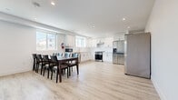 Located at 1297 1/2 W 36th St in Los Angeles, this modern three-story home offers seven bedrooms and seven bathrooms across three thoughtfully laid-out floors. The ground floor is the largest and serves as the communal hub, featuring an open-concept kitchen, dining room, and living room. The kitchen is outfitted with white shaker cabinetry, white subway tile backsplash, stainless steel appliances including a gas range and two full-sized refrigerators, and recessed lighting throughout. The adjacent dining area comfortably seats a large group and is bathed in natural light from wide windows. A cozy living room with a sectional sofa completes the shared space. Stainless steel stacked washer and dryer units are also conveniently located on this level. The upper two floors house the seven bedrooms, each bright and minimally styled with light wood-look flooring, recessed lighting, and windows offering neighborhood views. Bedrooms include desks, making them well-suited for focused work or study. The seven bathrooms are clean and contemporary, featuring white vanities with illuminated mirrors, and select bathrooms include a tub-shower combo with floor-to-ceiling white tile. A striking angled stairwell with clean white walls connects the floors, adding an architectural element to the interior.