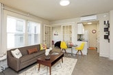 This single-floor unit offers a thoughtfully designed layout that balances openness with defined living spaces. The living area serves as the heart of the home, featuring large windows fitted with horizontal blinds that invite abundant natural light and frame views of the surrounding outdoors. A classic cast-iron radiator adds a charming, vintage character to the space, complementing the neutral color palette throughout.

The bedroom is separated from the main living area by elegant French doors, providing privacy while maintaining an airy, connected feel. Carpeted flooring, a wall-mounted mini-split heating and cooling unit, and a pendant light fixture complete this restful retreat.

The kitchen is efficiently arranged with white cabinetry, dark granite-style countertops offering ample prep space, and full appliance suite including a refrigerator, microwave, and range. A compact dining area adjacent to the kitchen provides a dedicated spot for meals.

The bathroom is bright and clean, featuring a window that brings in natural light, a pedestal-style vanity with under-cabinet storage, and a shower with curtain enclosure.

Throughout the unit, a cohesive modern-eclectic aesthetic is achieved through a consistent neutral base accented with bold pops of yellow and red, creating a vibrant yet inviting overall ambiance.